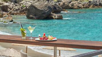 A wooden table with a plate of fruit and two drinks, overlooking a turquoise sea and a forested hillside.
