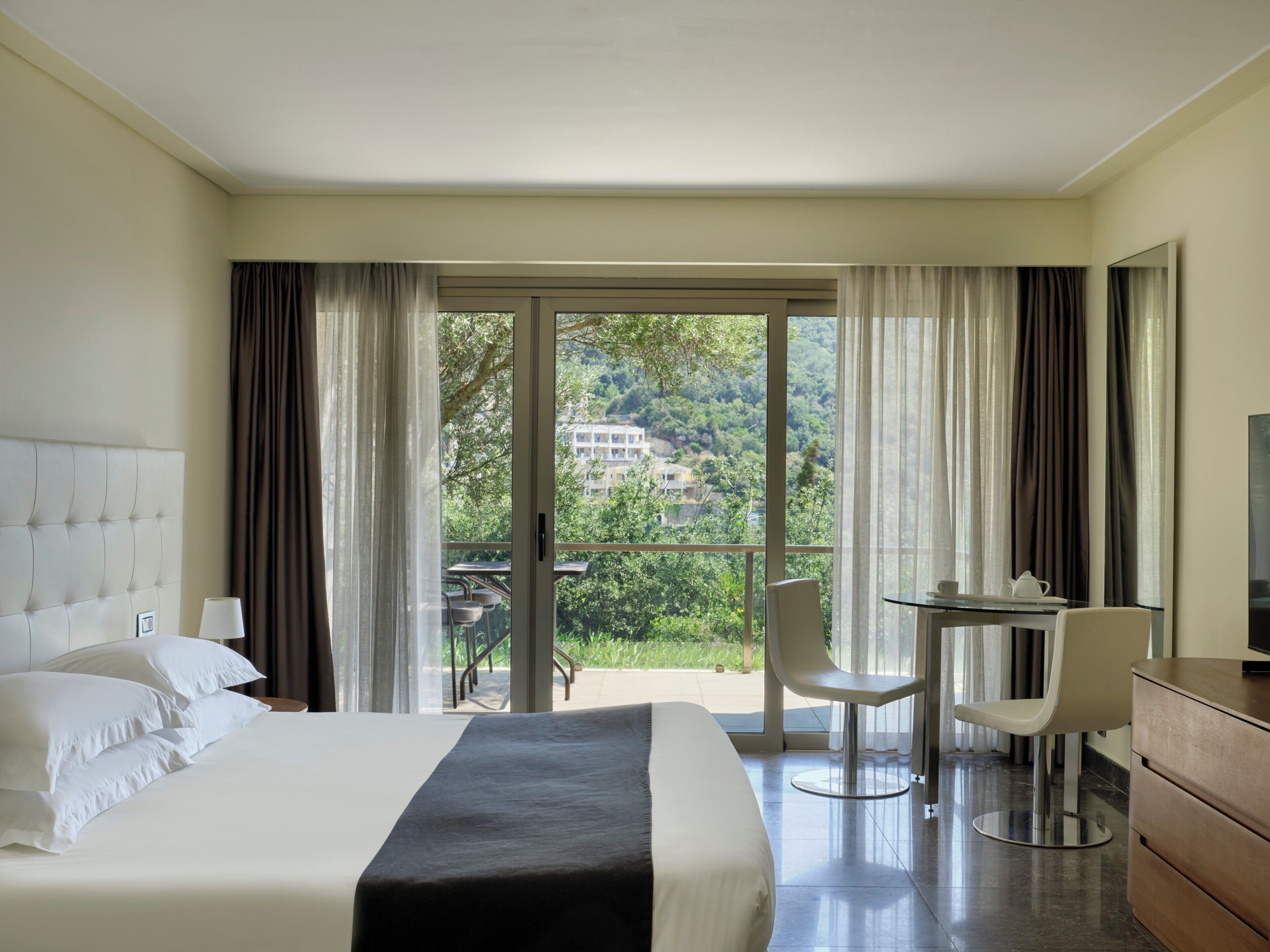 Habitación de hotel con cama, escritorio, dos sillas y un balcón con vistas a zonas verdes y edificios.