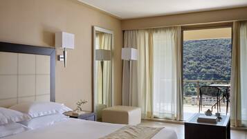 A hotel room with a large bed, a bedside table, a chair, a small table with a teapot, and a balcony with a view of a forested hillside.