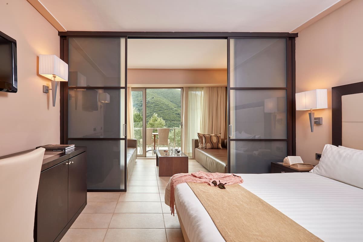 A hotel room with a bed, a desk, a chair, a sofa, a balcony with a view, and a television.