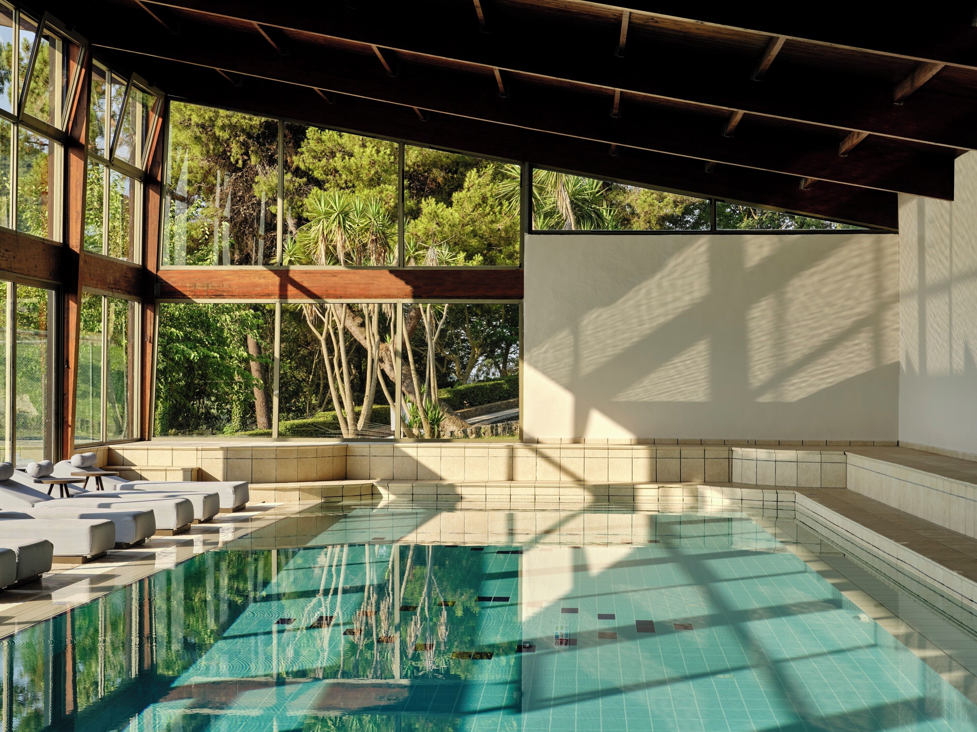A swimming pool with clear water, surrounded by lounge chairs and a view of trees through large windows.