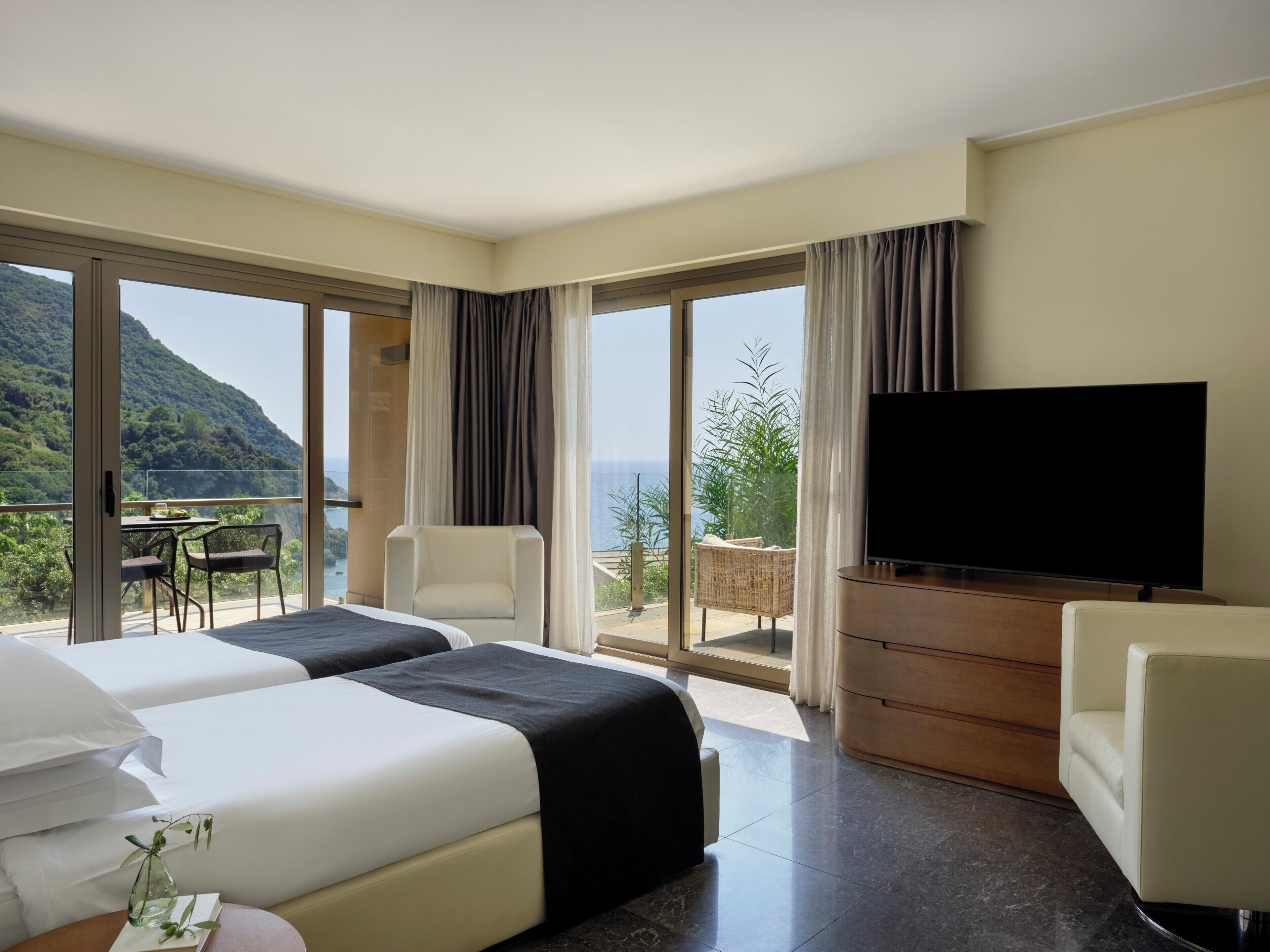 Une chambre d’hôtel avec un lit, une télévision, un balcon avec une table et des chaises, et une vue sur la mer.