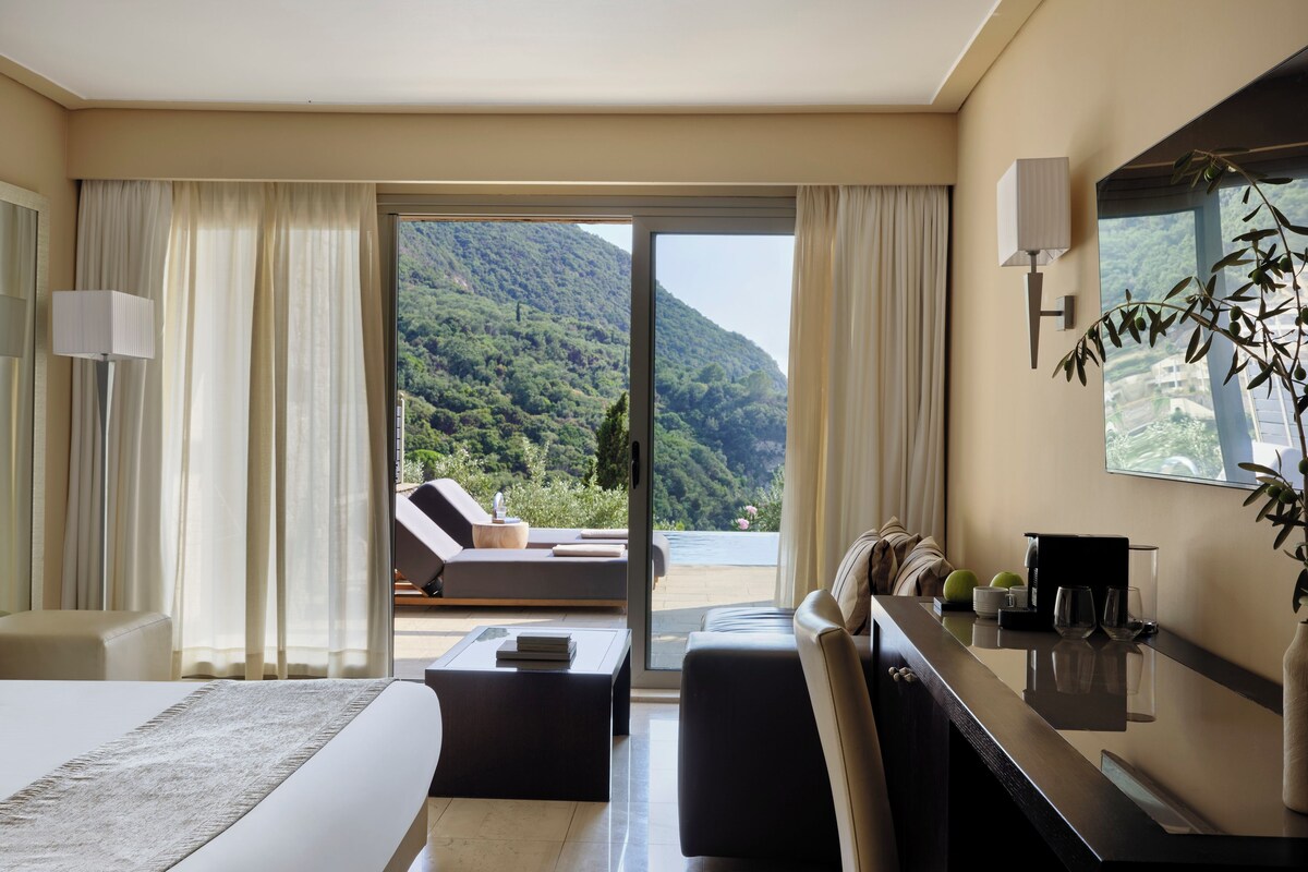 A hotel room with a bed, a desk, a chair, a sofa, a coffee table, and a view of a mountainous landscape.