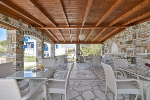 Un patio al aire libre con muros de piedra, vigas de madera y mobiliario de mimbre blanco.