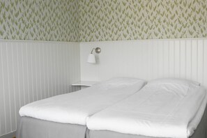 A neatly made bed in a room with a patterned wallpaper and a white bedside table with a lamp. - Pensionat Neptun (Bastad)