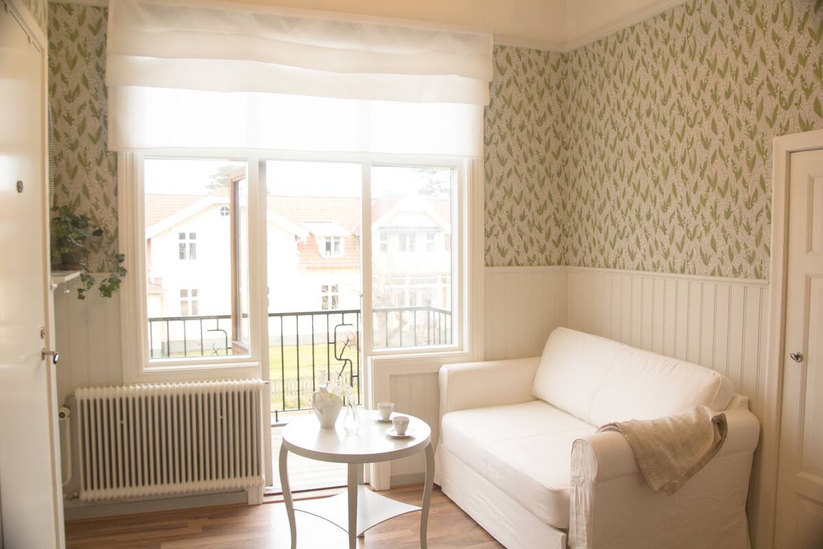Um quarto com um sofá branco, uma mesa redonda pequena com um vaso e chávenas, um radiador e uma janela com vista para casas.