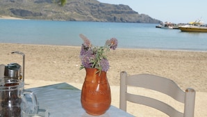 A table with a coffee maker and a vase of flowers, a chair, and a view of a beach with boats and a mountain in the background. - Costas & Chrysoula (Agios Vasileios)