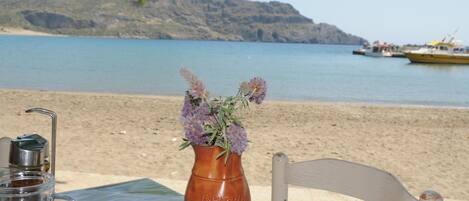 Uma mesa com uma cafeteira e um vaso de flores, uma cadeira e vista para uma praia com barcos e uma montanha ao fundo.