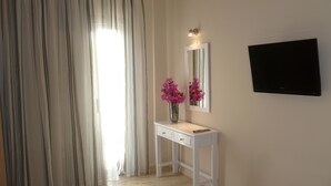 A room with a white vanity, a mirror, a vase with pink flowers, a flat-screen TV, and a large window with curtains. - Costas & Chrysoula (Agios Vasileios)