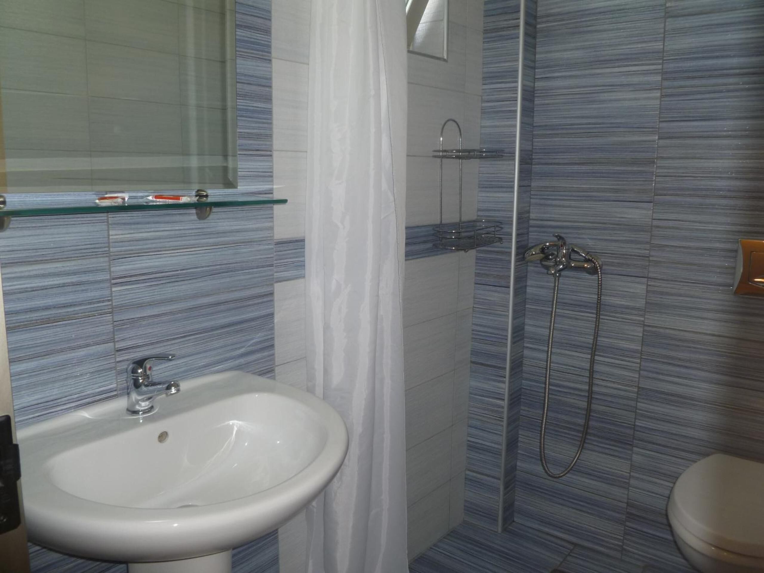 Un baño con lavamanos blanco, repisa de vidrio, ducha con cabezal de ducha manual y retrete.