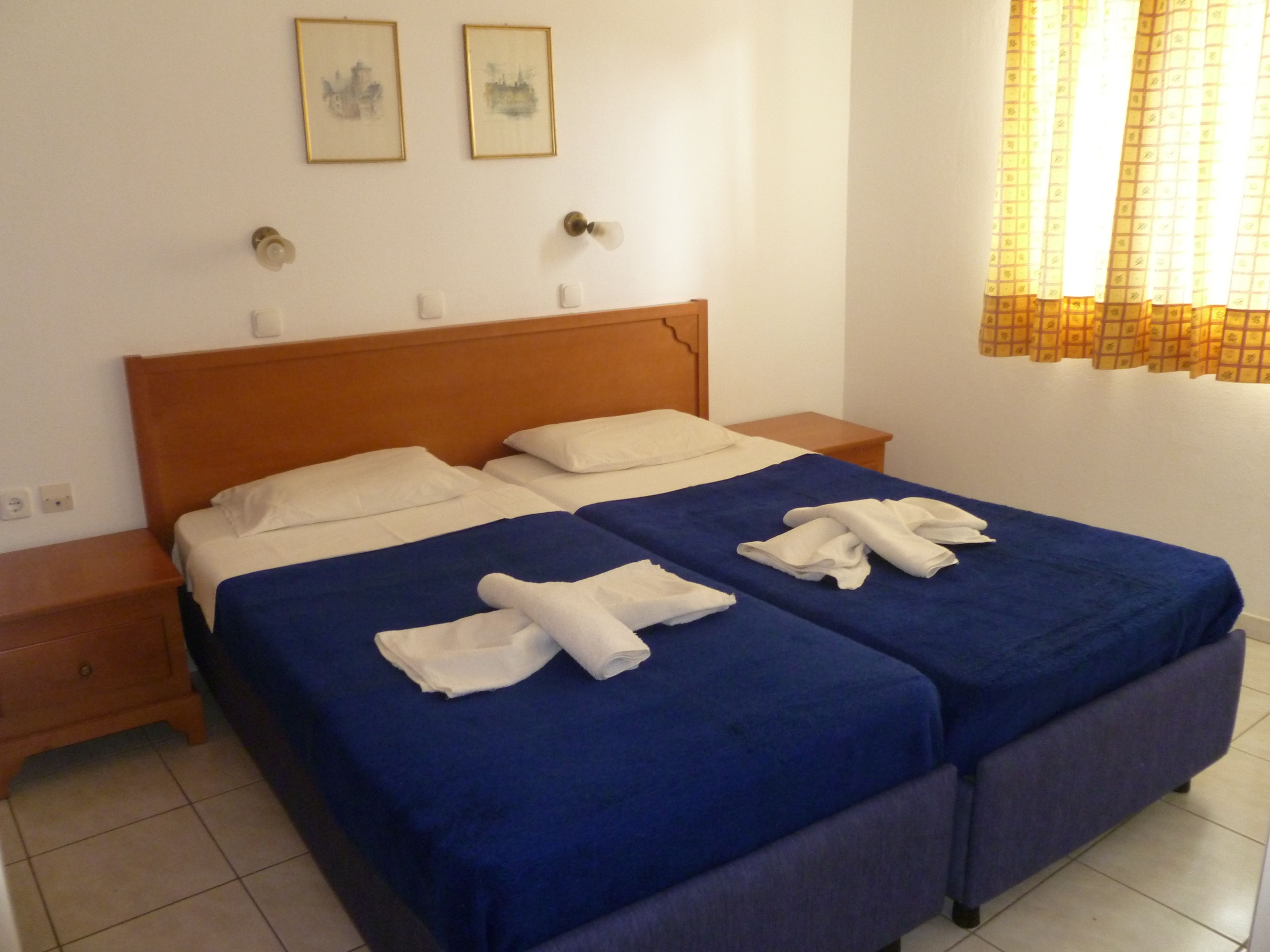 Cama doble con ropa de cama azul y toallas blancas, cabecero de madera, mesitas de noche y dos cuadros enmarcados en la pared.
