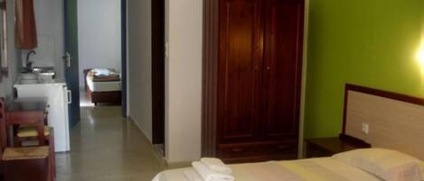 A hotel room with a bed, a wooden wardrobe, a small desk with a chair, and a kitchenette in the background.