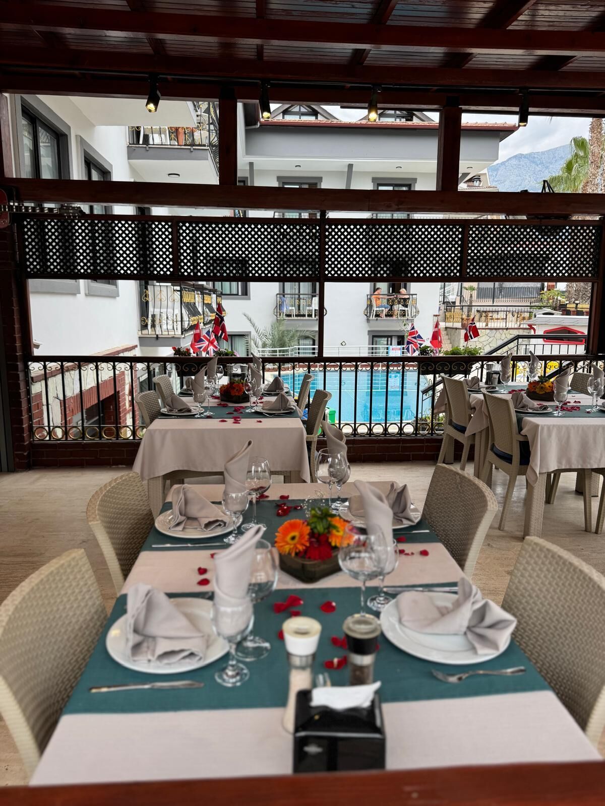 A restaurant with tables set for dining, a pool in the background, and a view of buildings and mountains.