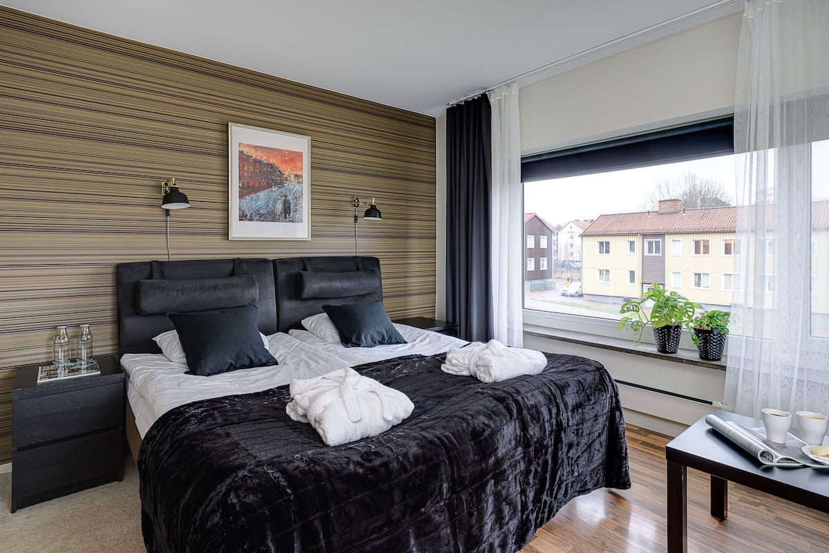 A modern hotel room with a large bed, two bedside lamps, a framed picture, and a view of buildings through the window.