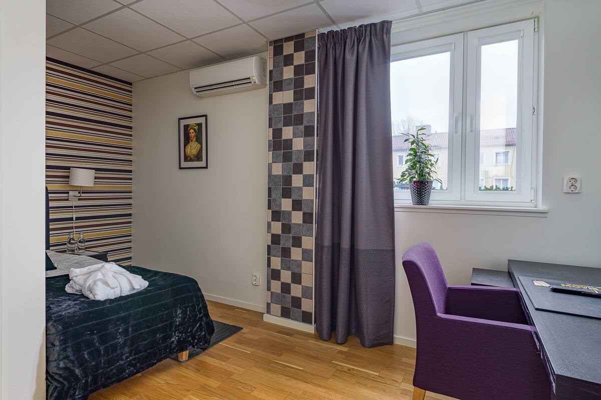 A room with a bed, a desk, a chair, a window with a curtain, a wall-mounted air conditioner, and a potted plant.