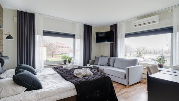 A modern hotel room with a bed, a grey sofa, a flat-screen TV, and large windows. - Wärdshuset C. Dickens (Fagersta)