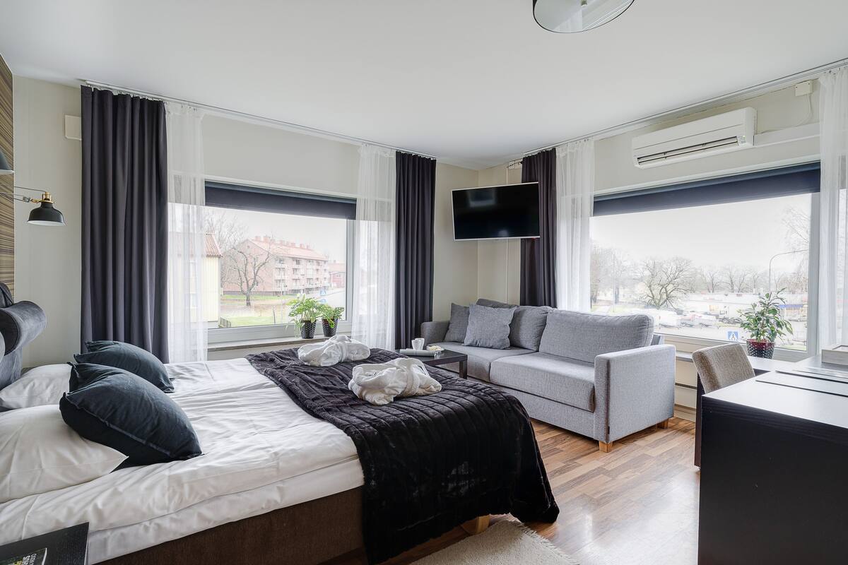 A modern hotel room with a bed, a grey sofa, a flat-screen TV, and large windows.