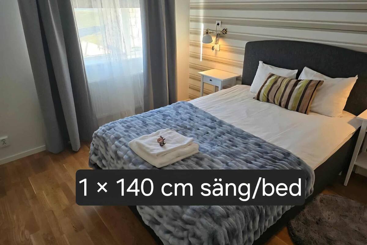 A hotel room with a bed measuring 140 cm in width, a grey duvet, and a small bedside table with a lamp.