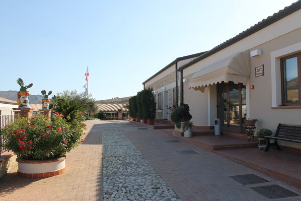 A paved walkway leading to a building with a reception area, potted plants, and a bench.