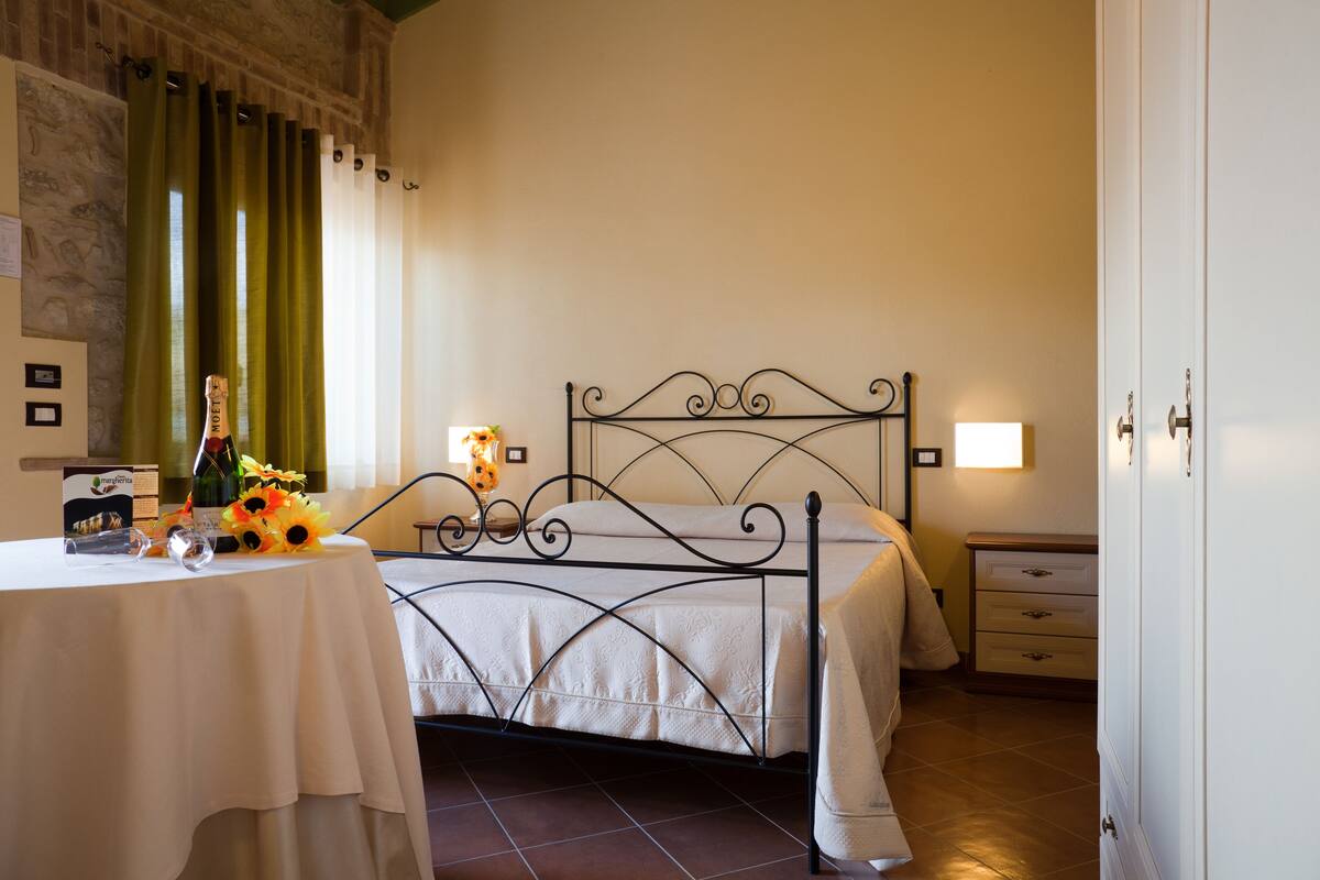 Un dormitorio ordenado con estructura metálica para la cama, una mesita de noche con una botella y flores, y un tocador con una lámpara.