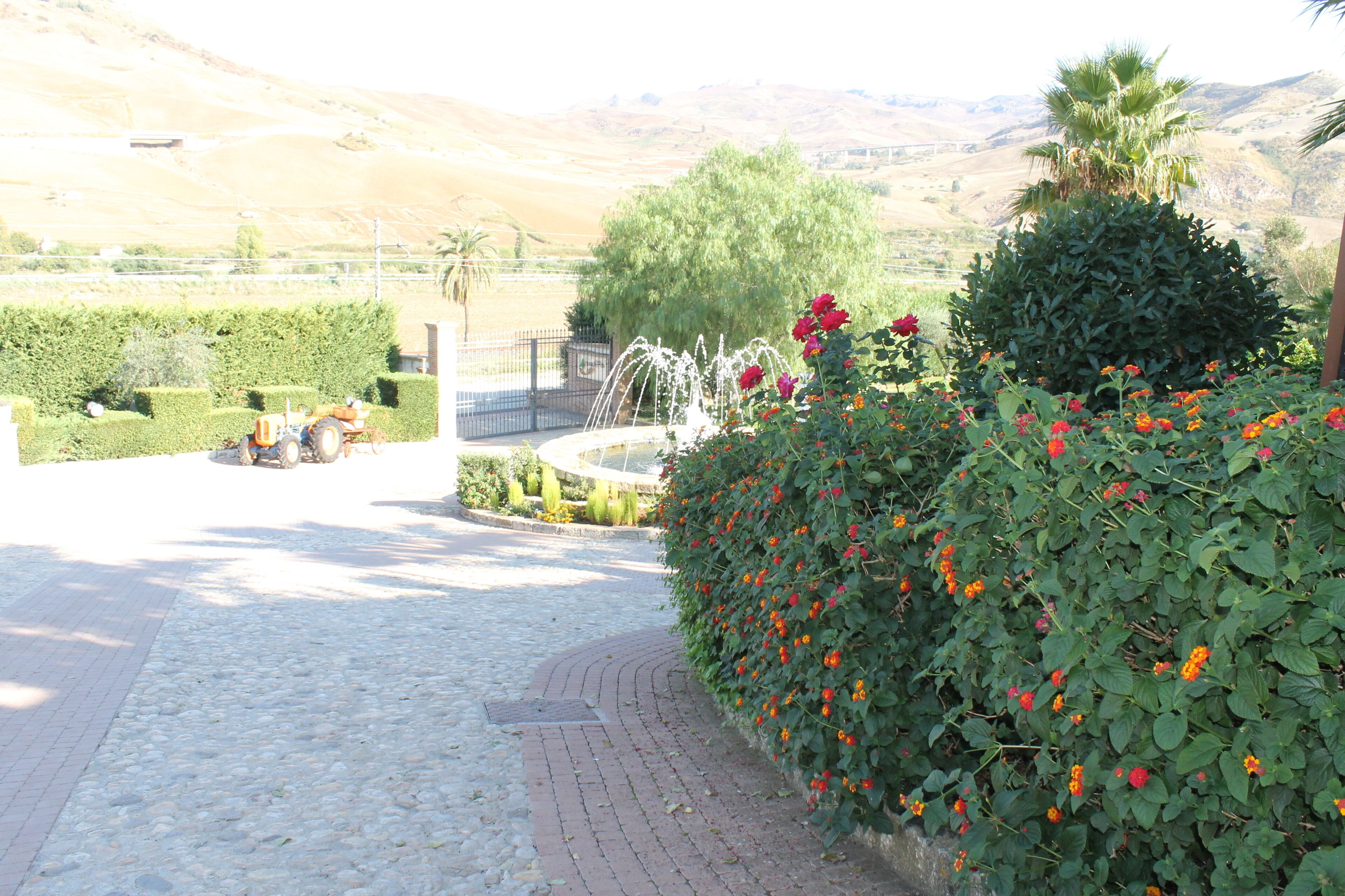 Un sendero pavimentado que conduce a un área ajardinada con una fuente, rodeada de setos y plantas con flores.