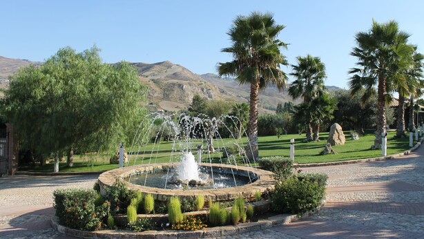 Un jardín ajardinado con una fuente circular, palmeras y montañas al fondo.