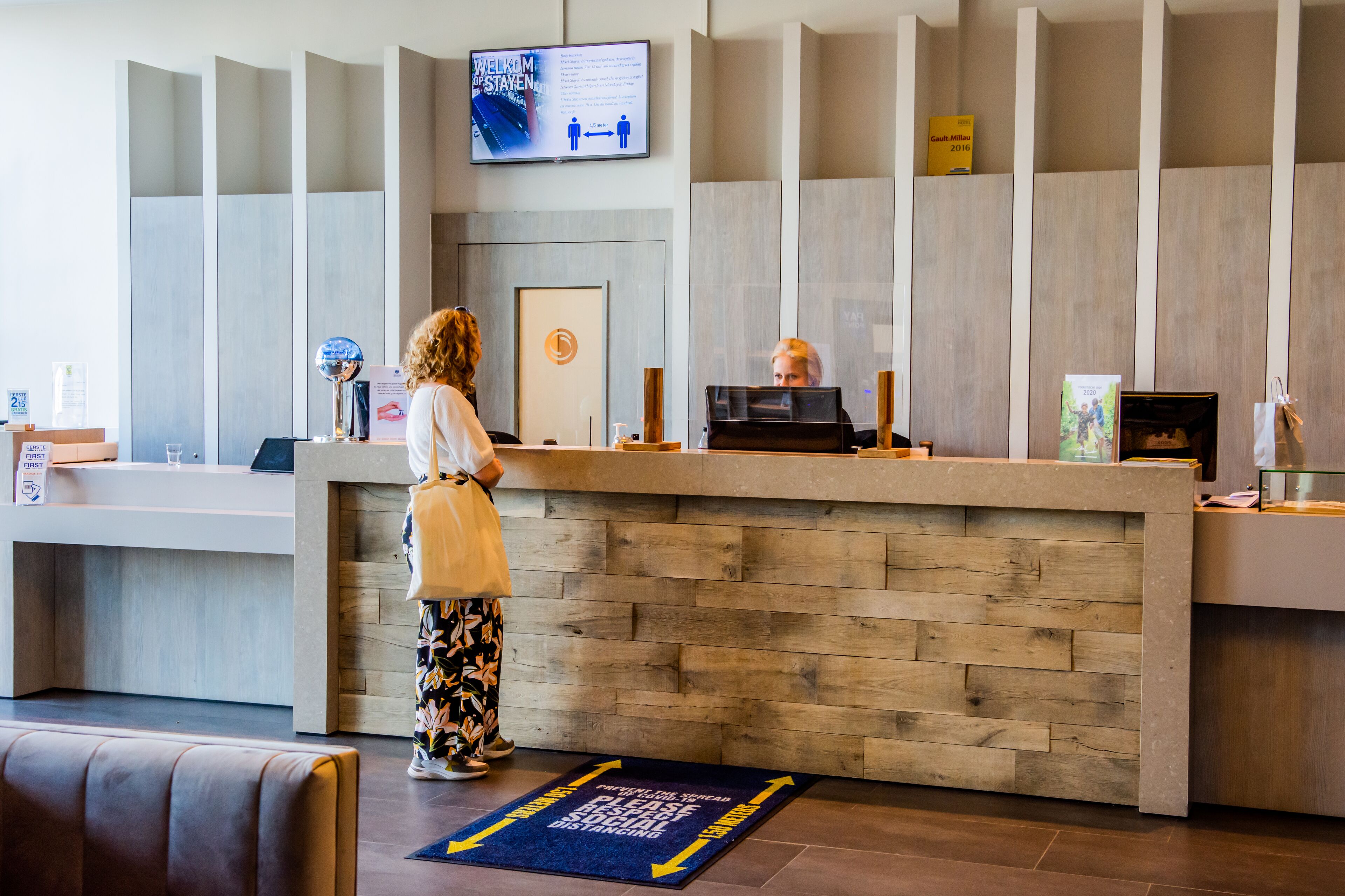 Un mostrador de recepción con paneles de madera, una pantalla digital en la parte superior y un tapete azul indicando la dirección en el suelo.