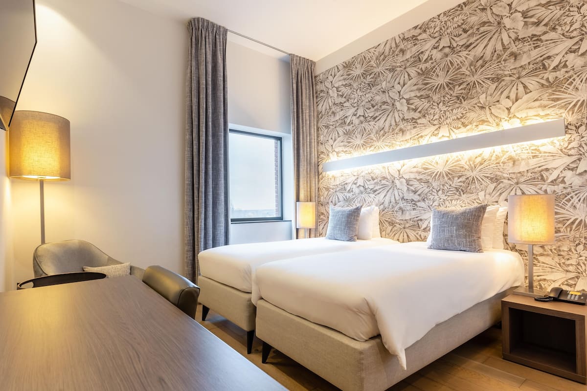 A hotel room with a large bed, a desk, two chairs, a desk lamp, a bedside table with a lamp, and a floral wallpaper.