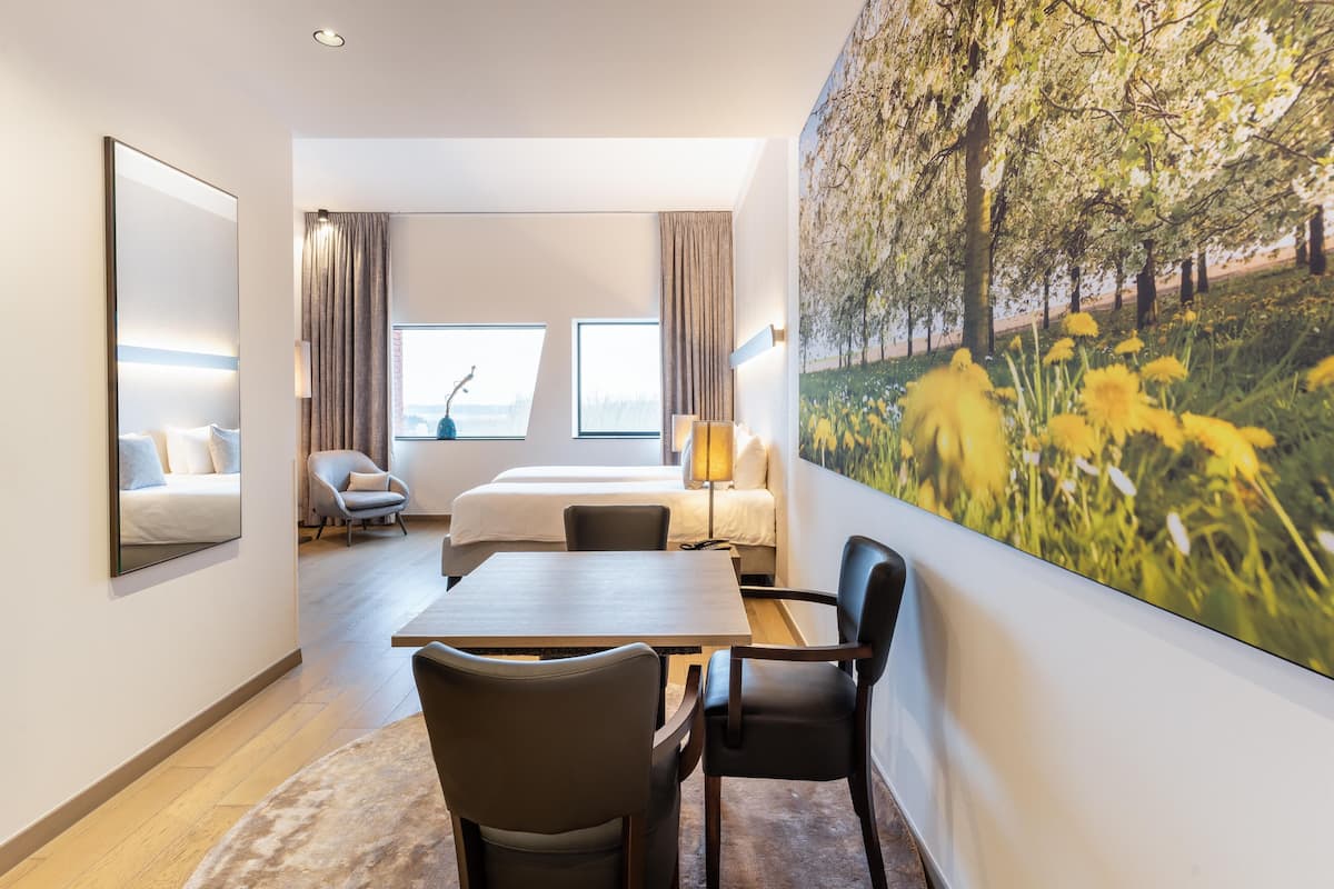 A hotel room with a bed, a desk, two chairs, a large window, a painting of a tree, and a mirror.