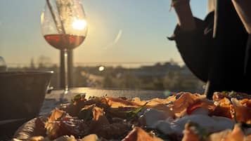 Une pizza sur une table, accompagnée d’un verre de vin rouge et d’une paille, le tout mis en valeur par une vue sur les gratte-ciel de la ville.