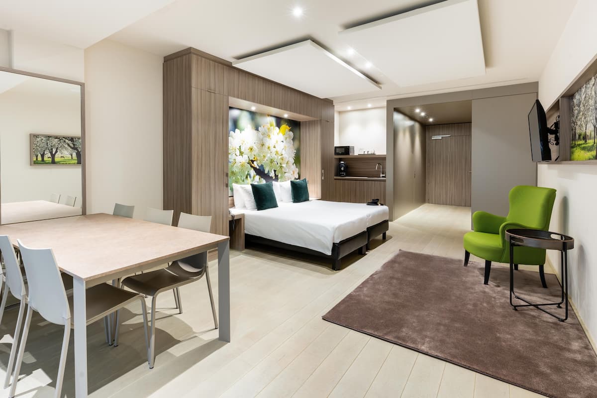 A modern hotel room with a bed, a dining table, and a green armchair.