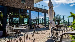 An outdoor café with tables and chairs, a large umbrella, and a building with a sign reading "CAFÉ DU SOLEIL".