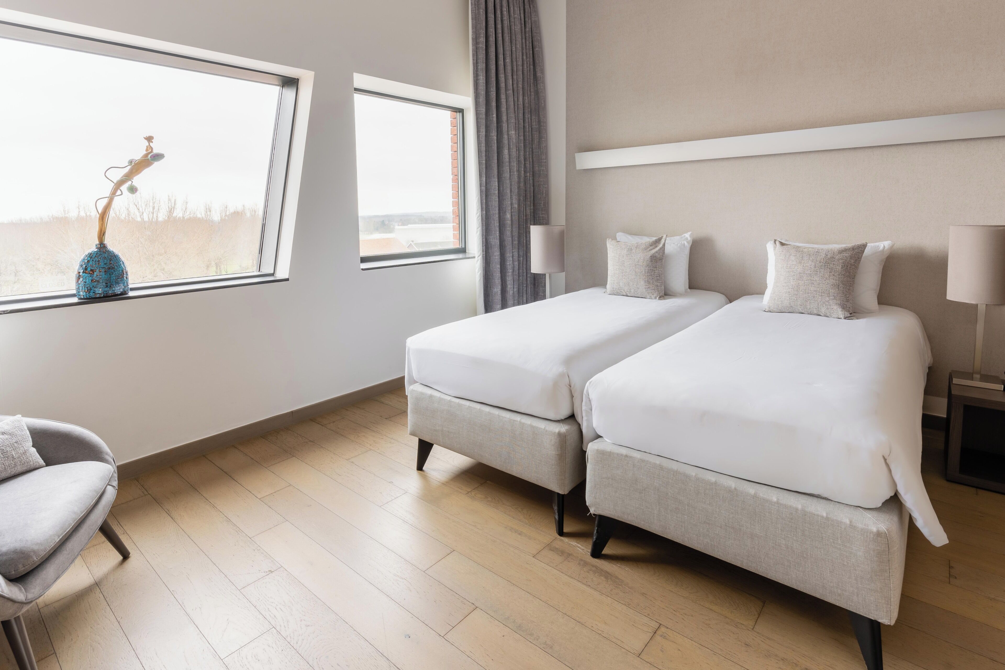 A hotel room with two beds, a chair, a lamp, and a window with a view.