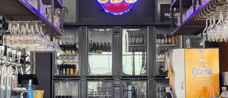 A bar area with a neon sign reading "CDS club drinks sport", shelves with glasses, and a refrigerator stocked with beverages.