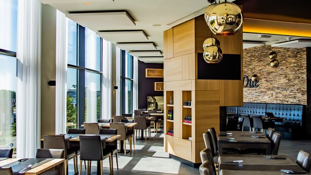 A modern hotel lobby with large windows, a central wooden reception desk, and multiple dining areas with tables and chairs.