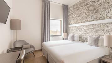 Quarto de hotel com cama, escrivaninha, cadeira, televisão e janela com cortinas.