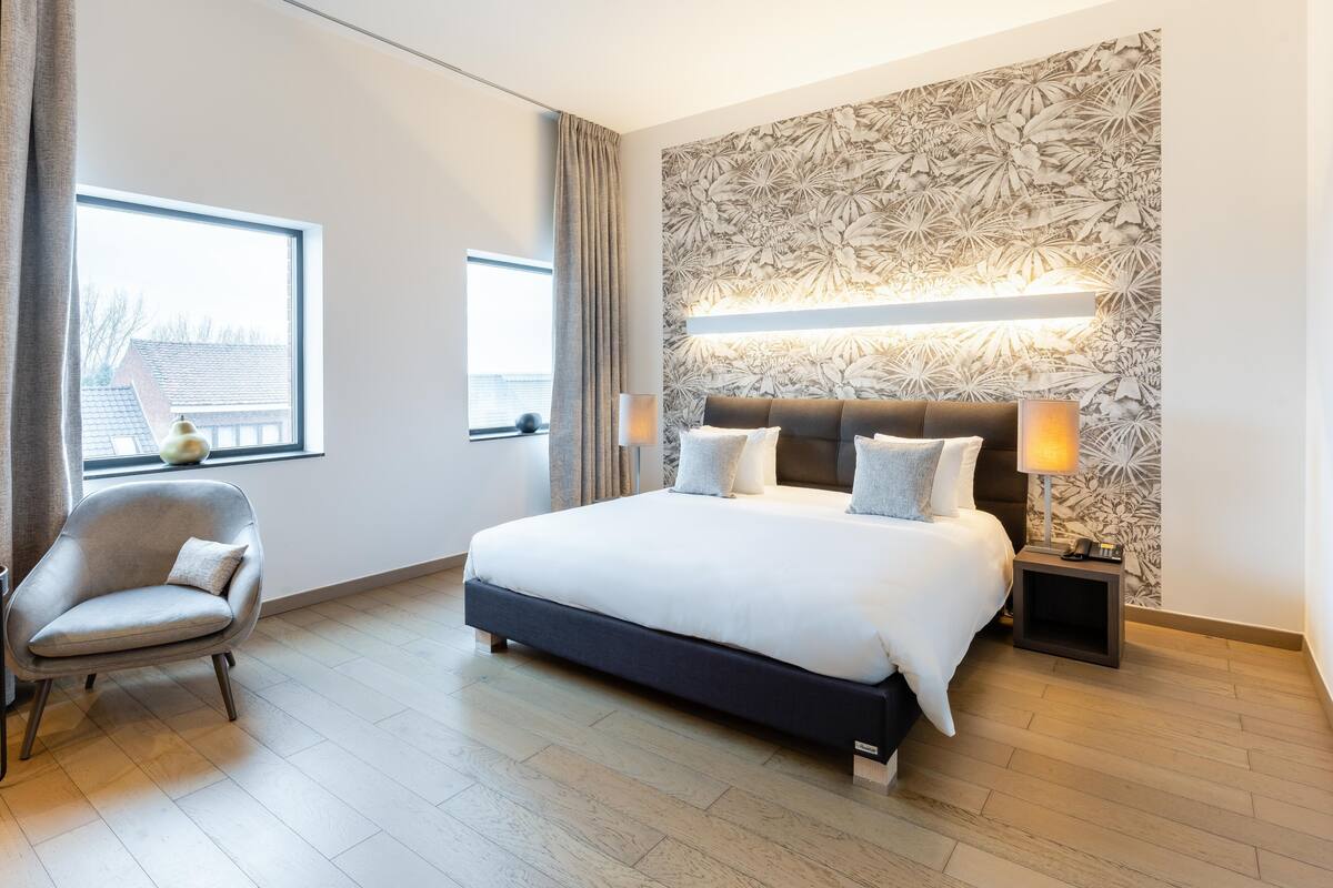 A modern bedroom with a large bed, two bedside tables with lamps, a chair, and a window with curtains.
