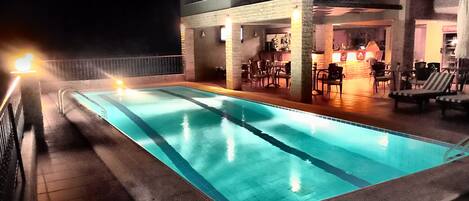 A rectangular outdoor swimming pool with a tiled edge, surrounded by a deck area with chairs and tables.