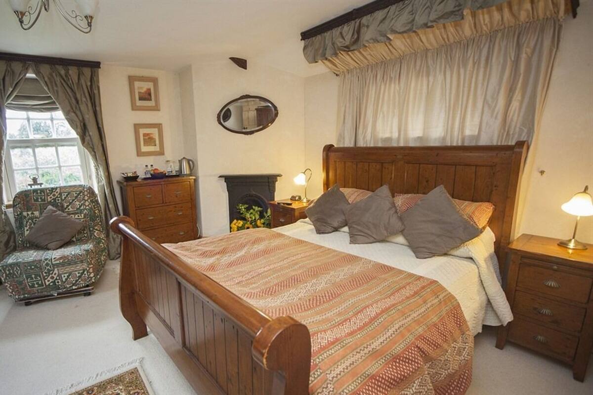 Une chambre à coucher avec un lit en bois, une commode, une chaise, une cheminée et une fenêtre avec des rideaux.
