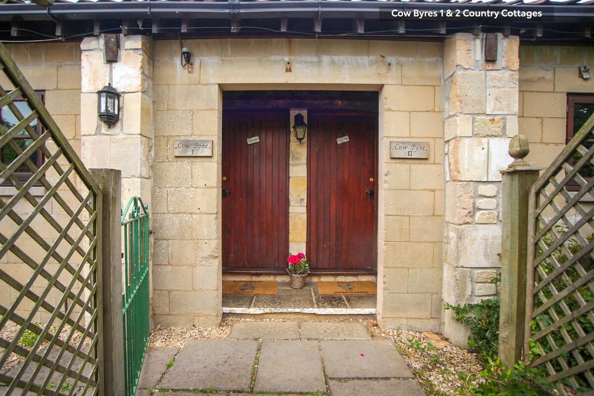 Une entrée traditionnelle avec deux portes en bois portant l’inscription « Cow Byres 1 et 2, maisons de campagne », un petit pot de fleurs et un mur en pierre.