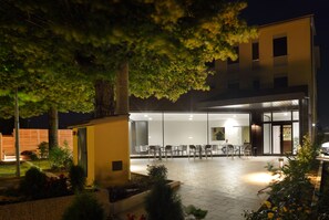 An outdoor dining area with tables and chairs, surrounded by greenery and a building with large windows. - Hotel Ristorante Dante (Torgiano)
