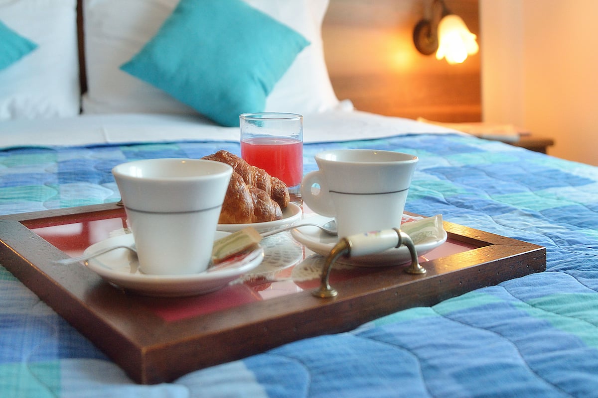 A breakfast tray with two cups of tea, a glass of juice, and a croissant on a bed with a blue quilt.