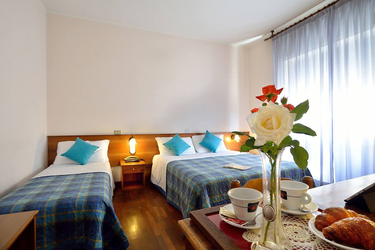 A hotel room with two beds, a wooden headboard, a bedside table with a lamp, a vase with flowers, a tray with croissants and a cup, and a window with curtains.