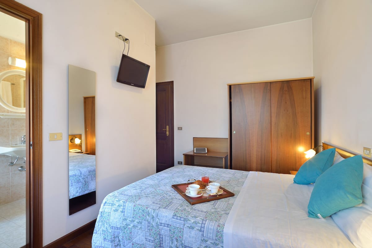A hotel room with a bed, a bedside table with a lamp, a wooden wardrobe, a mirror, and a bathroom visible through an open door.