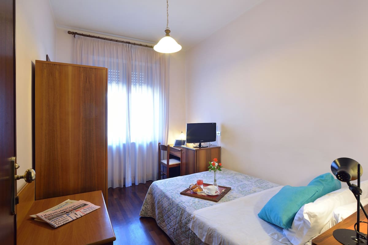 A hotel room with a bed, a desk with a computer, a chair, a television, and a tray with breakfast items.