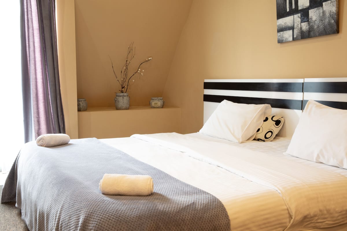 Ein ordentlich bezogenes Bett mit einem gestreiften Kopfteil, ein Nachttisch mit einer Vase gefüllt mit getrockneten Ästen und ein Fenster mit Vorhängen.