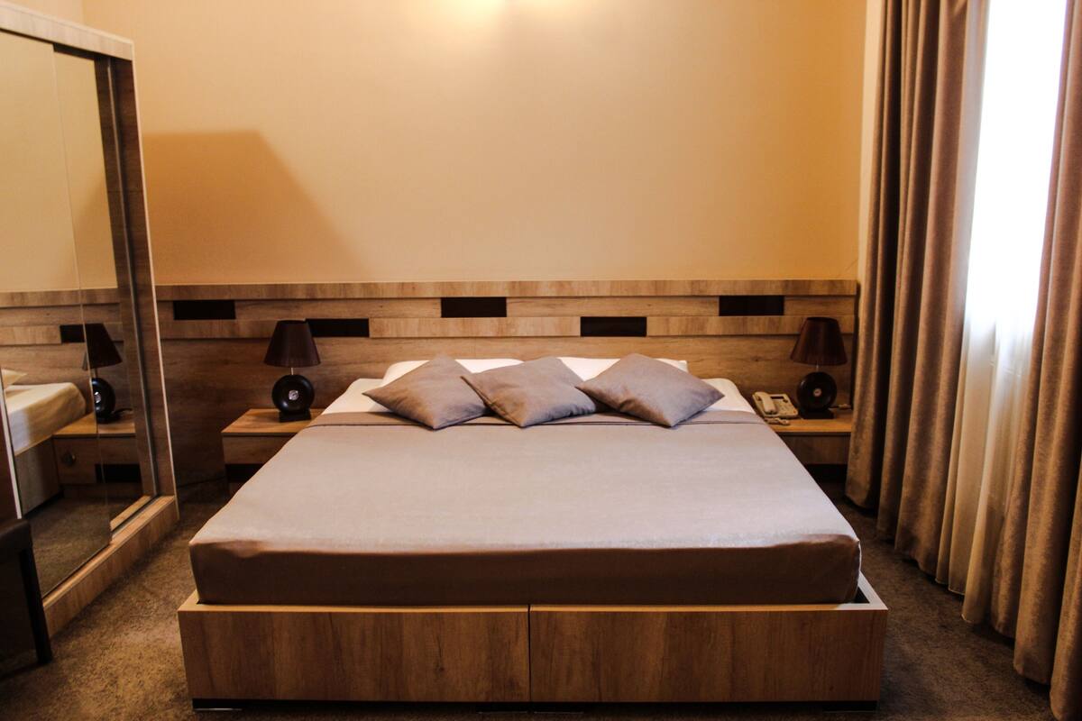 Ein Hotelzimmer mit einem großen Bett, einem Holzkopfende, Nachttischen mit Lampen, einem Spiegel und einem Fenster mit Vorhängen.