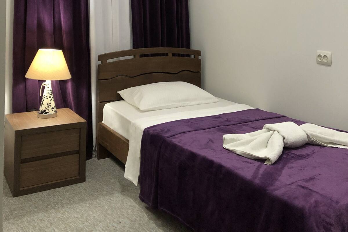 Ein Schlafzimmer mit einem Bett, einem Nachttisch mit Lampe, violetten Vorhängen und einem weißen Fensterrollo.