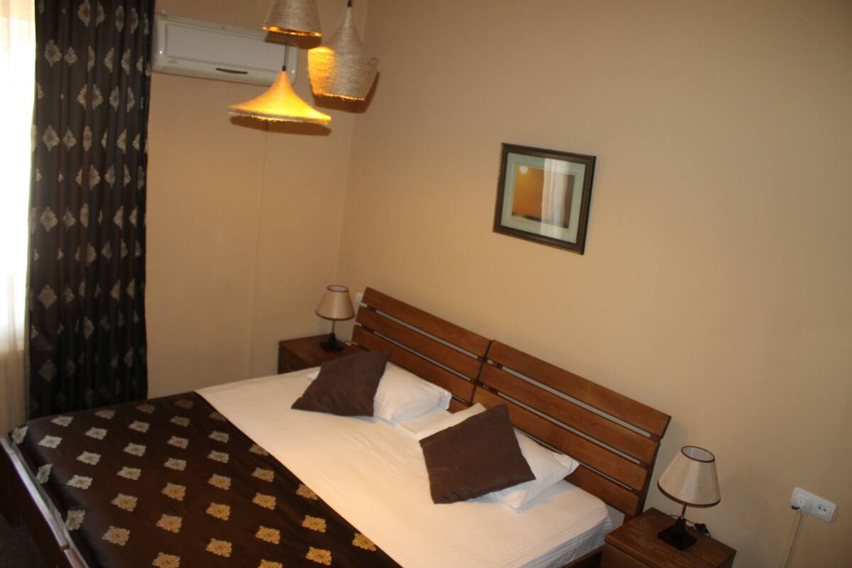 Ein ordentlich bezogenes Bett mit einer gemusterten Tagesdecke, zwei Nachttischlampen und einem gerahmten Bild an der Wand.