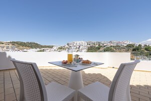 Una terraza con dos sillas de mimbre, una mesa con alimentos para el desayuno y vistas a un pueblo costero.